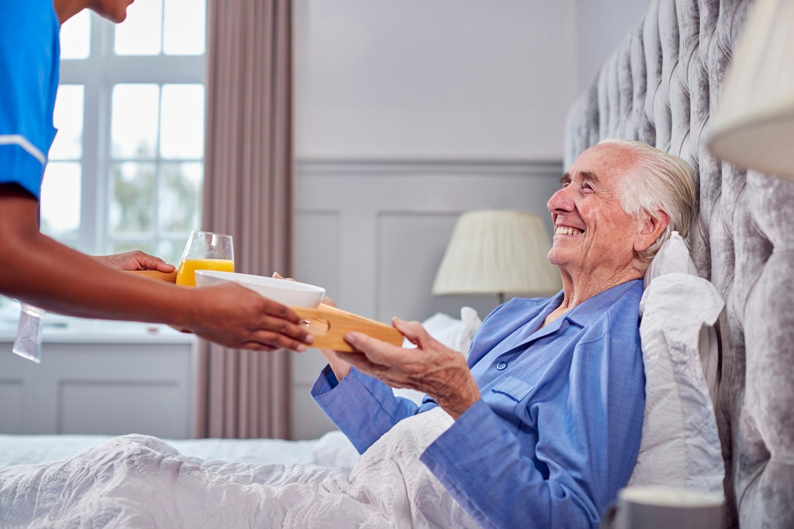 Female Care Worker In Uniform Bringing Senior Man At Home Breakfast In Bed On Tray