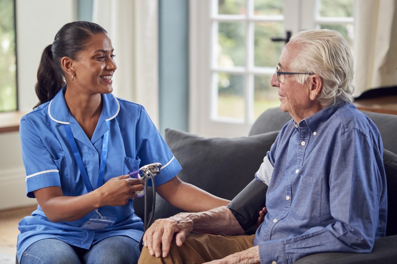 Old man in the lounge has his blood pressure taken by a female urgent home care worker.